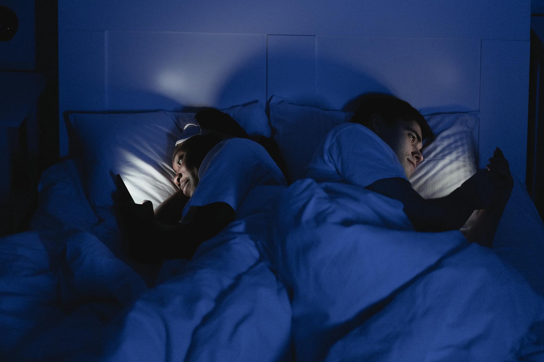 man and woman lying back to back in bed using smartphone